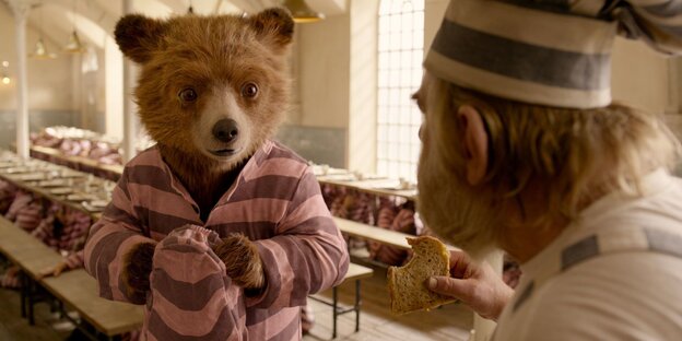 Im rosagestreiften Anzug steht Paddington vor einem Fließband. Im rosagestreiften Anzug steht Paddington vor einem Fließband.