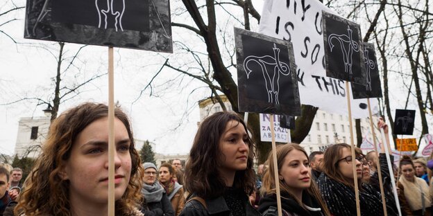 Frauen halten Schilder hoch, auf denen ein Uterus abgebildet ist, der symbolisch mit einem seiner Eileiter den Stinkefinger zeigt