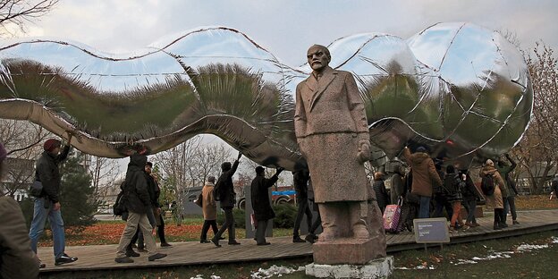 Menschen tragen eine riesige silberne Raupe über ihren Köpfen, im Vordergrund: eine Lenin-Statue Menschen tragen eine riesige silberne Raupe über ihren Köpfen, im Vordergrund: eine Lenin-Statue