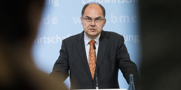 Ein Mann mit Halbglatze und in Anzug steht an einem Rednerpult mit Mikrofon Ein Mann mit Halbglatze und in Anzug steht an einem Rednerpult mit Mikrofon