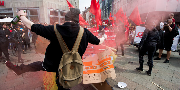 Bei einer Demo tritt ein vermummter Mann mit Bier in der Hand einen Gegenstand weg