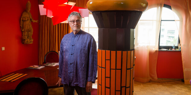 John Dew steht in seinem Wohnzimmer, die Wände bunt, neben ihm eine bunte Säule John Dew steht in seinem Wohnzimmer, die Wände bunt, neben ihm eine bunte Säule