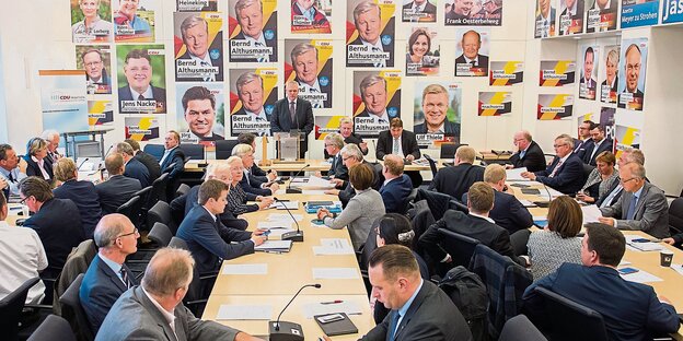 Blick in einen Raum mit vielen Männern, an den Wänden hängen Plakate von noch mehr Männern Blick in einen Raum mit vielen Männern, an den Wänden hängen Plakate von noch mehr Männern