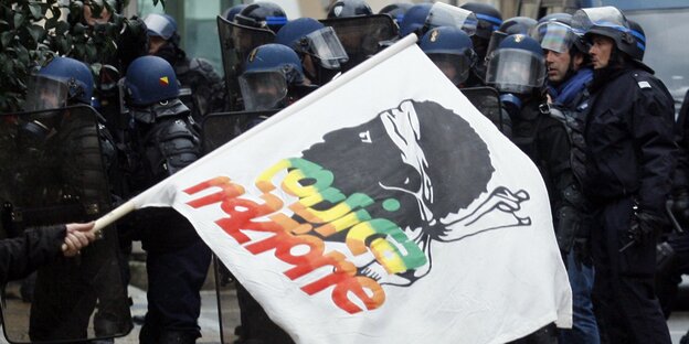 Eine Mann schwenkt eine korsische Fahne vor behelmten Polizisten Eine Mann schwenkt eine korsische Fahne vor behelmten Polizisten