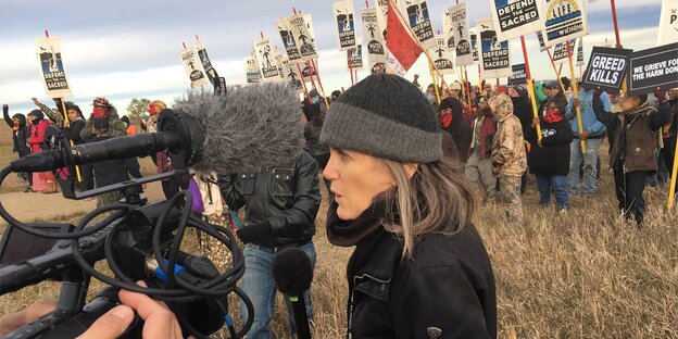 Eine Frau umgeben von Mikros auf einem Feld, im Hintergrund eine Demonstration