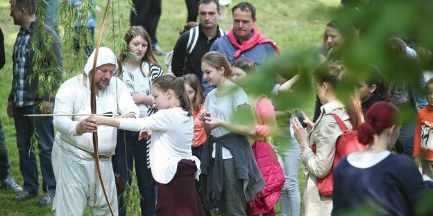 Ein kostümierter Mann leitet ein Mädchen im Bogenschießen an - hinter ihr eine Schlange weiterer Mädchen Ein kostümierter Mann leitet ein Mädchen im Bogenschießen an - hinter ihr eine Schlange weiterer Mädchen
