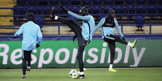 Drei Männer mit Mützen und hellblauen Trainingsjacken machen Dehnübungen auf dem grünen Rasen Drei Männer mit Mützen und hellblauen Trainingsjacken machen Dehnübungen auf dem grünen Rasen