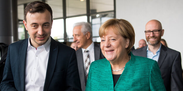 Ein junger Mann in Hemd und Sakko neben einer Frau mit orangenen Haaren und grünem Blazer