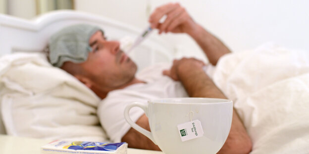 Ein Mann liegt mit Krankenhaushaube im Bett, ein Thermometer im Mund, eine Tasse Tee auf dem Nachttisch Ein Mann liegt mit Krankenhaushaube im Bett, ein Thermometer im Mund, eine Tasse Tee auf dem Nachttisch