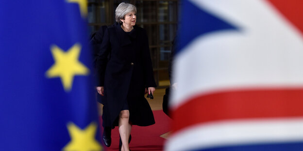 Eine Frau im schwarzen Mantel läuft auf einem roten Teppich, am Rand des Bilds sieht man eine europäische und eine britische Flagge Eine Frau im schwarzen Mantel läuft auf einem roten Teppich, am Rand des Bilds sieht man eine europäische und eine britische Flagge