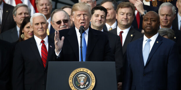 Donald Trump steht an einem Rednerpult und spricht in ein Mikrofon, neben ihm stehen mehrere Menschen Donald Trump steht an einem Rednerpult und spricht in ein Mikrofon, neben ihm stehen mehrere Menschen