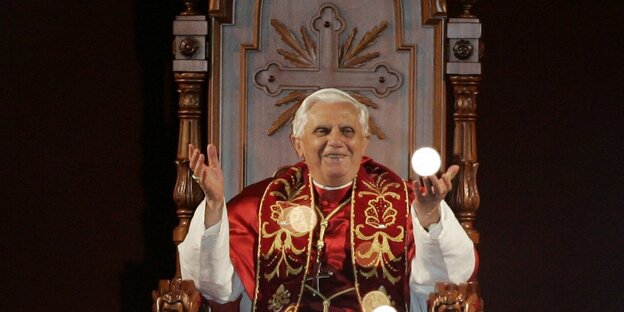 Ex-Papst Benedixt XVI. sitzend, die Hände in die Höhe haltend Ex-Papst Benedixt XVI. sitzend, die Hände in die Höhe haltend