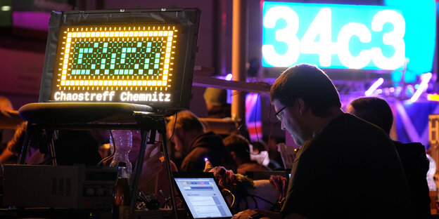 Menschen sitzen in einer dunklen Halle vor hell leuchtenden Wegweisern und blau schimmernden Laptops Menschen sitzen in einer dunklen Halle vor hell leuchtenden Wegweisern und blau schimmernden Laptops