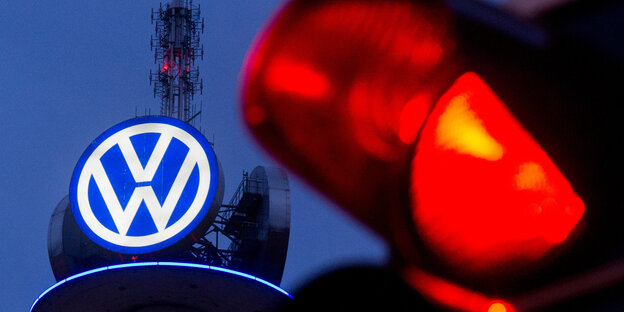 Ein großes Logo von Volkswagen leuchtet in der Dämmerung, davor ist eine rote Ampel zu sehen Ein großes Logo von Volkswagen leuchtet in der Dämmerung, davor ist eine rote Ampel zu sehen