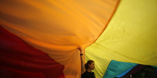 Eine Frau steht unter einer großen Regenbogenfahne und stützt sie mit ihrem Arm Eine Frau steht unter einer großen Regenbogenfahne und stützt sie mit ihrem Arm
