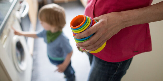 Ein Kind steht an einer Waschmaschine, eine Mutter hält Kinderspielzeug in der Hand