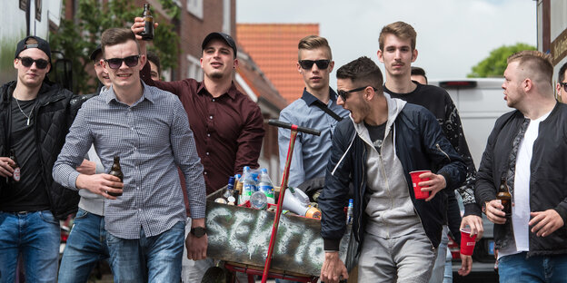 Junge Männer trinken aus Bechern und ziehen einen Wagen hinter sich her Junge Männer trinken aus Bechern und ziehen einen Wagen hinter sich her