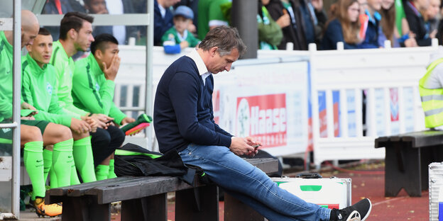 Ein mittelalter Mann sitzt auf einer Bank vor Fußball-Spielern und Fans und schaut auf sein Handy. Ein mittelalter Mann sitzt auf einer Bank vor Fußball-Spielern und Fans und schaut auf sein Handy.
