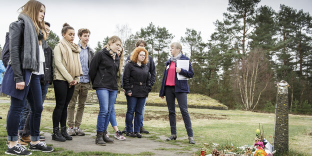 Eine Gruppe von Jugendlichen steht vor einem Gedenkstein und hört einer älteren Frau zu, die ein Namensschild und einen Stapel Papier trägt. Eine Gruppe von Jugendlichen steht vor einem Gedenkstein und hört einer älteren Frau zu, die ein Namensschild und einen Stapel Papier trägt.