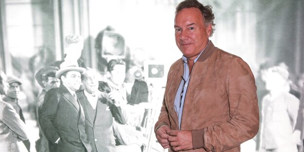 Nico Hofmann steht in brauner Jacke vor dem Still eines schwarz-weiß-Films Nico Hofmann steht in brauner Jacke vor dem Still eines schwarz-weiß-Films