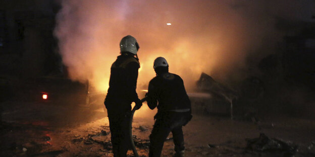 Zwei Männer, die Schutzhelme tragen, richten in der Dunkelheit einen Schlauch auf ein Feuer Zwei Männer, die Schutzhelme tragen, richten in der Dunkelheit einen Schlauch auf ein Feuer