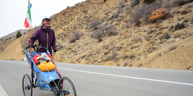 Kristine Paltén läuft mit einem Handwagen, an dem die iranische und die schwedische Fahne wehen, über eine Asphaltstraße durch die Wüste Kristine Paltén läuft mit einem Handwagen, an dem die iranische und die schwedische Fahne wehen, über eine Asphaltstraße durch die Wüste