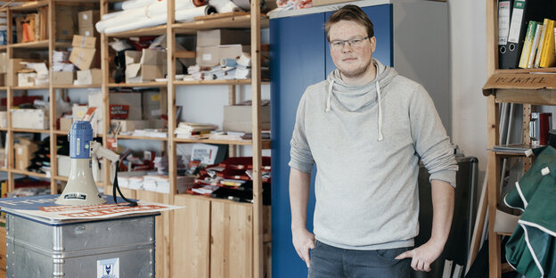 Ein junger Mann in grauem Pullover, im Hintergrund Holzregale, eine blaue Tür, neben ihm ein Schrank, auf dem ein Megafon ist Ein junger Mann in grauem Pullover, im Hintergrund Holzregale, eine blaue Tür, neben ihm ein Schrank, auf dem ein Megafon ist