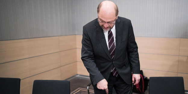 Ein Mann in Anzug setzt sich auf einen Stuhl und zeigt nach unten Ein Mann in Anzug setzt sich auf einen Stuhl und zeigt nach unten