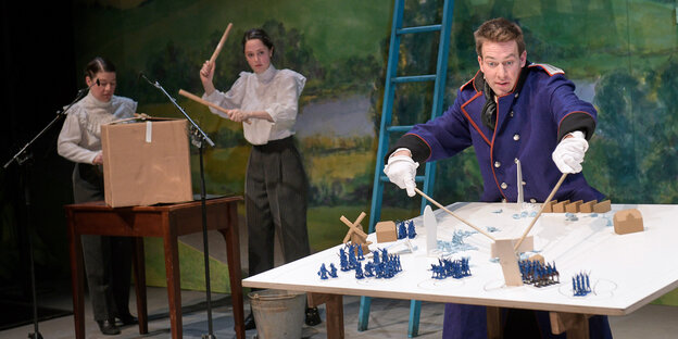 Zwei Schauspielerinnen in Biedermeierblusen haben Instrumente zum Lärmen in der Hand, ein Schauspieler in Uniform verschiebt kleine Soldaten. Zwei Schauspielerinnen in Biedermeierblusen haben Instrumente zum Lärmen in der Hand, ein Schauspieler in Uniform verschiebt kleine Soldaten.