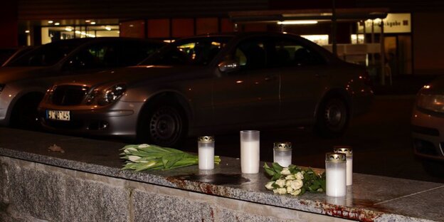 Kerzen und Blumen auf einem Mäuerchen, dahinter parkende Autos, dahinter ein Supermarkt Kerzen und Blumen auf einem Mäuerchen, dahinter parkende Autos, dahinter ein Supermarkt