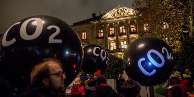 Menschen mit schwarzen Luftballons, auf denen „CO2“ steht vor einem erleuchteten Gebäude Menschen mit schwarzen Luftballons, auf denen „CO2“ steht vor einem erleuchteten Gebäude