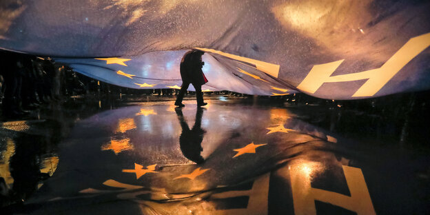 Ein Mensch verschwindet unter einer großen, blauen EU-Fahne, die sich über den Asphalt wölbt Ein Mensch verschwindet unter einer großen, blauen EU-Fahne, die sich über den Asphalt wölbt