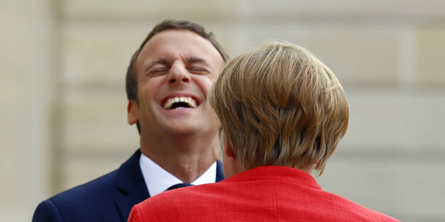 Macron lacht mit zurückgelegtem Kopf, Merkel steht davor Macron lacht mit zurückgelegtem Kopf, Merkel steht davor
