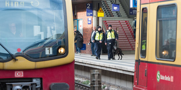 Zwei Sicherheitskräfte der Deutschen Bahn gehen in gelben Warnwesten mit einem Schäferhund über das Gleis, im Vordergrund fährt die Ringbahn. Zwei Sicherheitskräfte der Deutschen Bahn gehen in gelben Warnwesten mit einem Schäferhund über das Gleis, im Vordergrund fährt die Ringbahn.