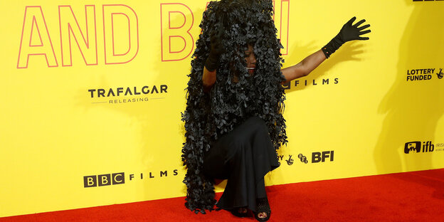 Grace Jones sitzt in einem schwarzen Kleid vor einer gelben Wand und streckt einen Arm aus Grace Jones sitzt in einem schwarzen Kleid vor einer gelben Wand und streckt einen Arm aus