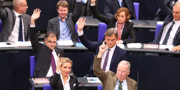 Viele Männer und zwei Frauen sitzen in Reihen hintereinander und heben die Hand Viele Männer und zwei Frauen sitzen in Reihen hintereinander und heben die Hand