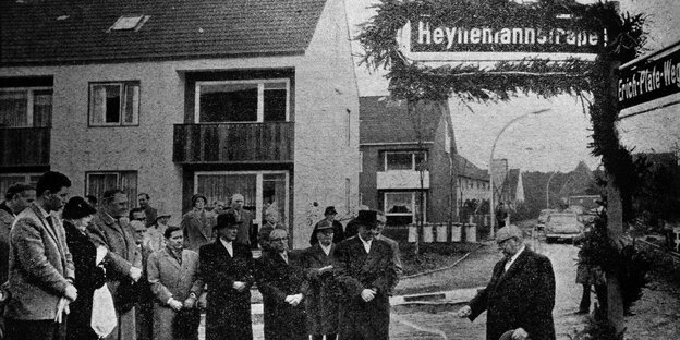 Ein alter glatzköpfiger Mann schneidet ein Band durch das an einem Straßenschild hängt. Auf dem Schild steht "Heynemannstraße".