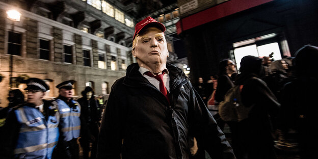 Jemand läuft mit einer Trumpmaske durch eine Stadt, im hintergrund stehen Polizisten Jemand läuft mit einer Trumpmaske durch eine Stadt, im hintergrund stehen Polizisten