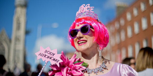 Eine Frau trägt eine pinkfarbene Perücke, Sonnenbrille und ein Krönchen. Sie hält ein Schild „Body Autonomy“ in die Höhe Eine Frau trägt eine pinkfarbene Perücke, Sonnenbrille und ein Krönchen. Sie hält ein Schild „Body Autonomy“ in die Höhe