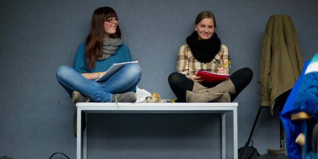 Zwei junge Frauen sitzen im Schneidersitz auf einem Tisch und halten Blöcke zum Mitschreiben. Zwei junge Frauen sitzen im Schneidersitz auf einem Tisch und halten Blöcke zum Mitschreiben.
