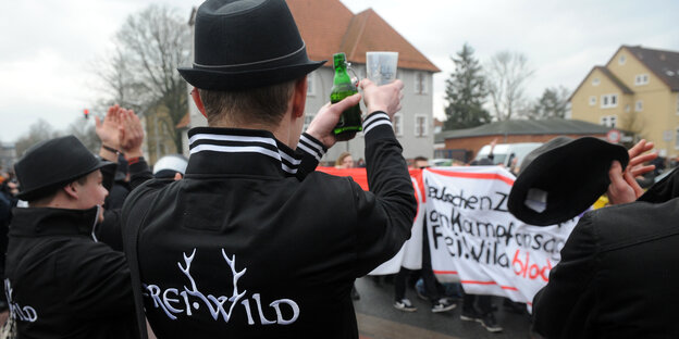Drei Leute in schwarzen Kleidern mit dem Schriftzug "Freiwild" stehen vor einer Menschenmenge, die gegen das Konzert der Band demonstriert. Drei Leute in schwarzen Kleidern mit dem Schriftzug "Freiwild" stehen vor einer Menschenmenge, die gegen das Konzert der Band demonstriert.