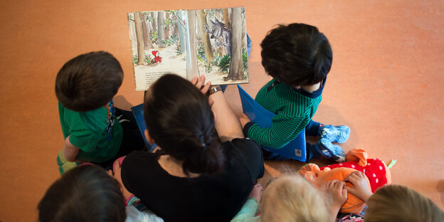 Eine Frau mit dunklen Haaren hält ein Bilderbuch in der Hand. Um sie herum sitzen Kinder und schauen auf das Buch.