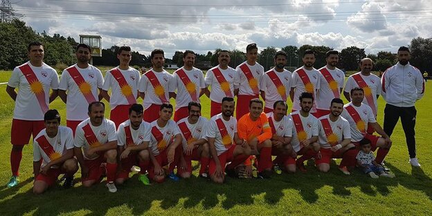 22 Spieler und ein Trainer der Mannschaft des FC Exidxan. Die Spieler tragen rot-weiße Trikots mit einer Sonne vorne drauf. 22 Spieler und ein Trainer der Mannschaft des FC Exidxan. Die Spieler tragen rot-weiße Trikots mit einer Sonne vorne drauf.