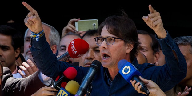 Mann mit Brille spricht mit erhobenen Händern vor Mikrofonen, im Hintergrund stehen Menschen Mann mit Brille spricht mit erhobenen Händern vor Mikrofonen, im Hintergrund stehen Menschen
