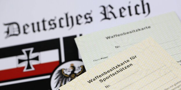 Ein Blatt Papier mit einer Abbildung der Fahne des Deutschen Reiches und des Eisernen Kreuzes. Auf dem Blatt liegen zwei Waffenscheine. Ein Blatt Papier mit einer Abbildung der Fahne des Deutschen Reiches und des Eisernen Kreuzes. Auf dem Blatt liegen zwei Waffenscheine.