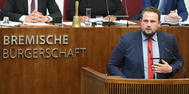 Ein junger Mann mit roter Krawatte und Anzug steht am Rednerpult der Bremischen Bürgerschaft und spricht. Ein junger Mann mit roter Krawatte und Anzug steht am Rednerpult der Bremischen Bürgerschaft und spricht.