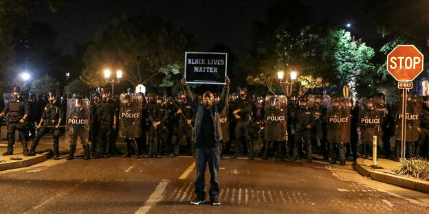 Ein schwarzer Mann steht vor einer Reihe von Polizisten mit einem Schild "Black Lives Matter" Ein schwarzer Mann steht vor einer Reihe von Polizisten mit einem Schild "Black Lives Matter"