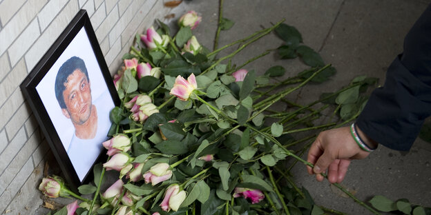 Ein Bild steht an eine Mauer gelehnt auf dem Boden und zeigt einen mittelalten Mann mit schwarzen Haaren. Vor dem Bild liegen Rosen.