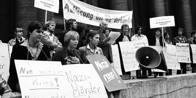 Mehrere Menschen sitzen und stehen an einer Mauer. Sie tragen Schilder um den Hals und halten Banner in die Höhe, auf denen sie die Veruteilung von Nazis und die Auflösung von SS-Organisationen fordern Mehrere Menschen sitzen und stehen an einer Mauer. Sie tragen Schilder um den Hals und halten Banner in die Höhe, auf denen sie die Veruteilung von Nazis und die Auflösung von SS-Organisationen fordern