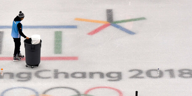 Eine Person schiebt eine Tonne über die Eisfläche an dem Logo der Olympischen Spiele vorbei Eine Person schiebt eine Tonne über die Eisfläche an dem Logo der Olympischen Spiele vorbei
